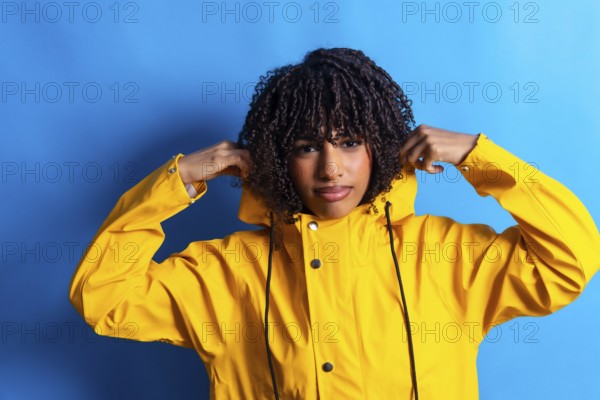 Studio shot of a young woman with curly hair adjusting the hood of her yellow raincoat against a vibrant blue backdrop