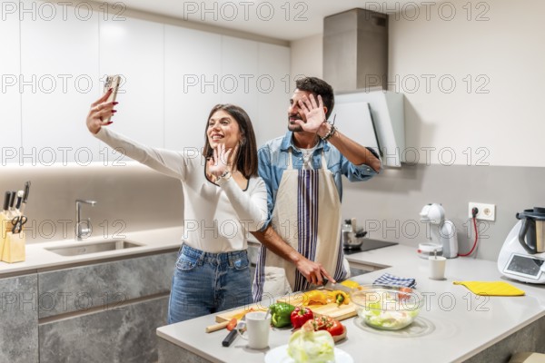 Young couple waving at the camera while preparing a healthy salad in their kitchen, making a video call with smartphone