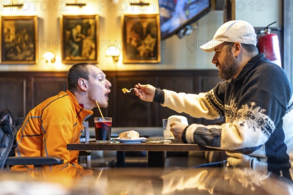 Man feeding his disabled friend in a restaurant, showcasing genuine care and inclusivity while sharing a joyful meal together