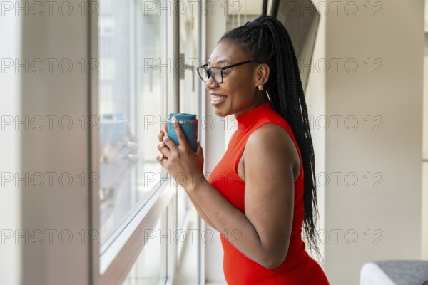 Smiling businesswoman drinking from a reusable cup while looking out the window of her hotel room during a business trip