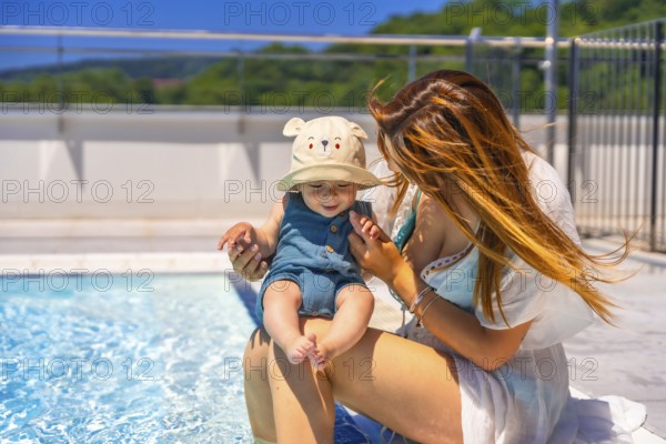 Adorable baby with funny hay and mother sitting on the edge of a pool in a sunny day of summer