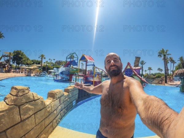 Bearded man enjoying a fun summer vacation at a water park, capturing joyful moments with a selfie on a sunny day by the pool
