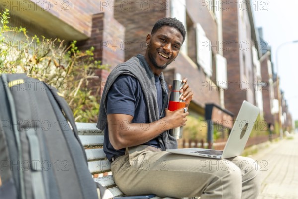 African businessman working with laptop while drinking from reusable bottle sitting on a street bench