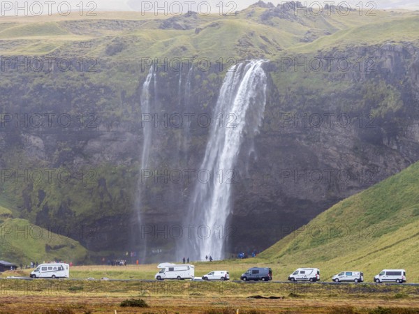 Tourist groups at waterfall Seljalandsfoss on a rainy day, queue at the path behind the waterfall, campervans, Iceland, Europe