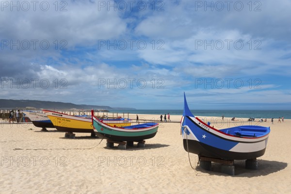 A row of colourful fishing boats on the sandy beach, some people walking along the shore, Praia da Nazare beach, Nazaré, Oeste, Leiria district, Centro, Estremadura, Portugal, Europe