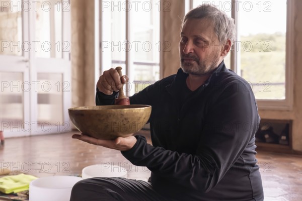 In a serene room, a mature man sits calmly, with a serene expression as he gently plays a bronze bowl. A moment of meditation