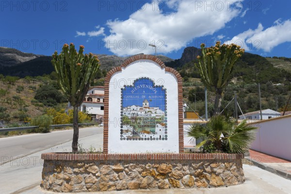 A welcome sign with colourful flowers and cacti in the foreground, surrounded by mountains under a cloudy sky, Canillas de Aceituno, Málaga province, Malaga, Andalusia, Spain, Europe
