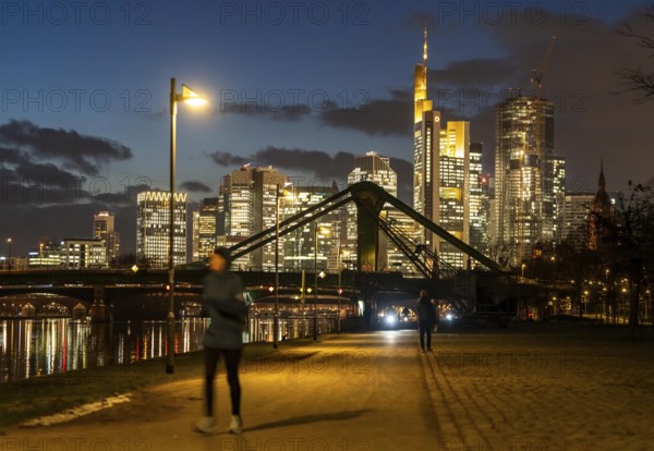 Skyline of the city centre of Frankfurt am Main, joggers on the pavement, promenade along the river Main, dusk, Hesse, Germany raft bridge in the background