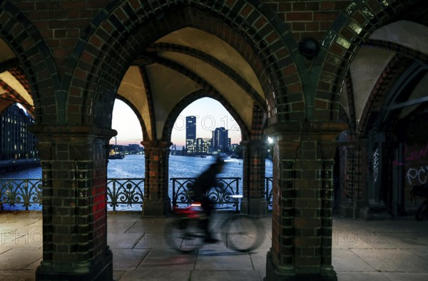 Bicycle rider to the sunrise at the Oberbaum Bridge, view through the arcades over the Spree to the Treptower Towers, Berlin, 17.10.2024, Berlin, Berlin, Germany, Europe