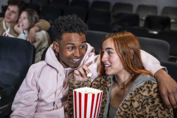 Happy diverse couple enjoying film and sharing popcorn bucket in movie theater, whispering during the projection
