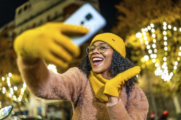 Smiling woman wearing yellow woolen gloves, hat and scarf taking selfie with her smartphone in a city street decorated with christmas lights