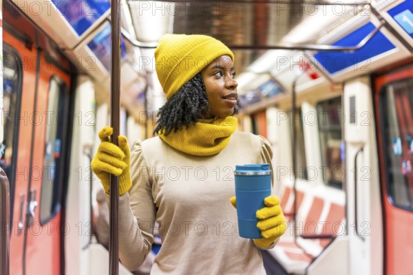 Black woman wearing yellow winter clothes holding a blue reusable coffee cup standing on a subway train
