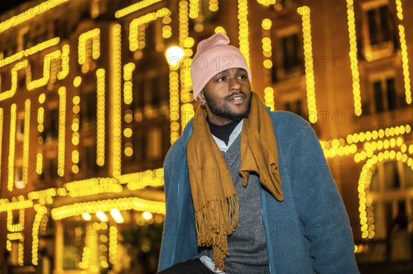African man sitting outdoors during Christmas night facing Christmas fairy lights decorating a facade