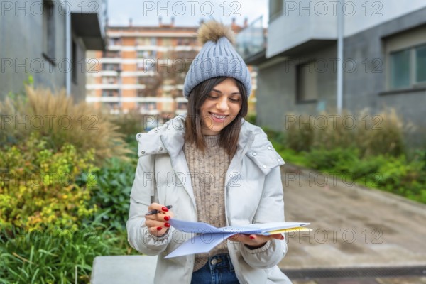 Smiling student writing notes in a notebook sitting on a city bench in winter