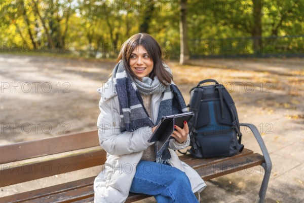 Woman using digital tablet sitting on a park bench during a sunny day of autumn