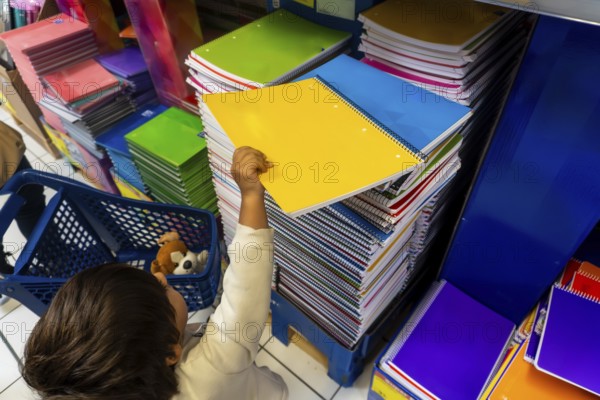 A child is reaching for a yellow spiral notebook in a store. The store is filled with many notebooks of different colors and sizes. Back to school concept