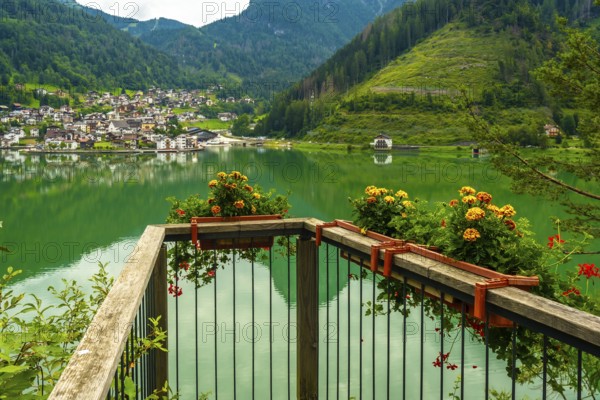 Wooden balcony with flower boxes overlooking the picturesque village of alleghe, reflecting on emerald green lake waters in the dolomites