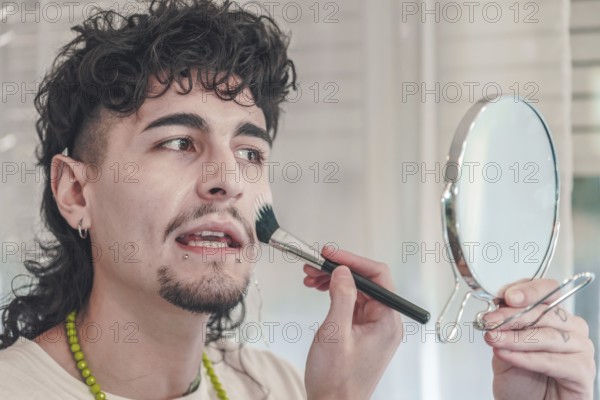 Young man with curly hair and piercings is applying makeup to his face using a brush while looking in a small mirror held in his hand, showcasing diversity and inclusion in beauty routines