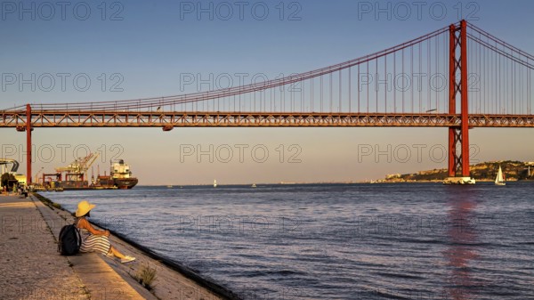 A person sits by a riverbank, gazing at the bridge and boats during a calm sunset, 25 de Abril Bridge, Lisbon