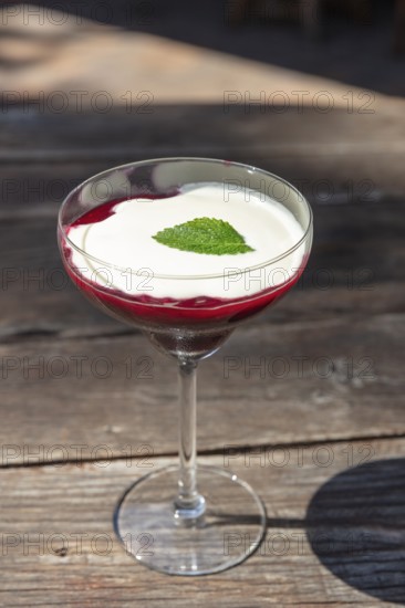 Elegant dessert in a glass with red raspberry sauce, white cream and mint leaf on a wooden table, red fruit jelly dessert
