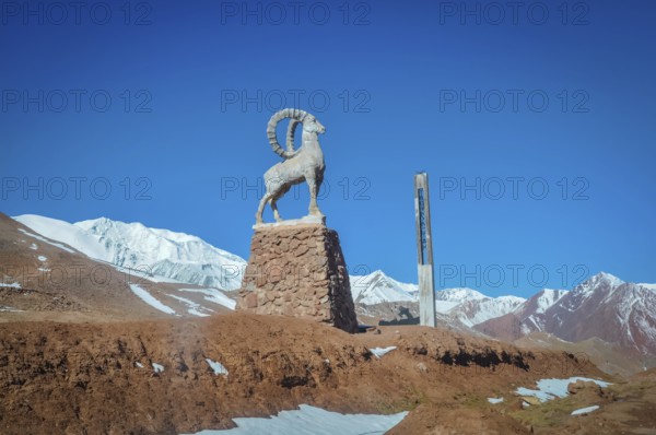 Sculpture of bouquetin on stony base surrounded by mountains with snowy peaks in distance in Karakul in Tajikistan