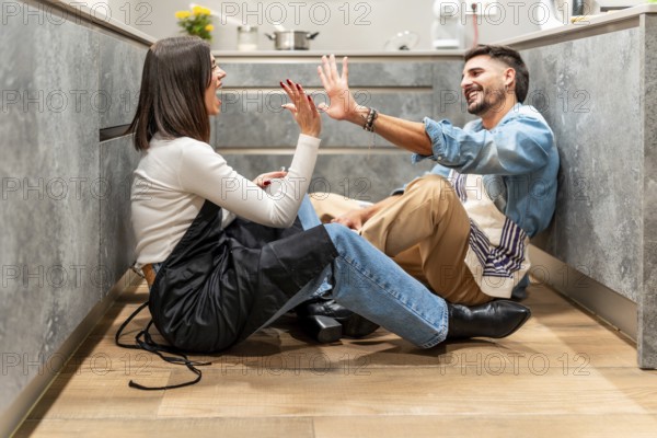 Two young chefs are sitting on the kitchen floor, giving each other a high five and laughing, celebrating their successful work