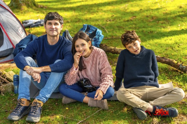 Happy caucasian father, daughter and son sitting next to camping tent enjoying nature in the forest during weekend