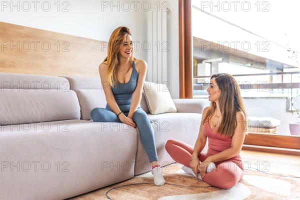 Two smiling women wearing yoga clothes are chatting and relaxing in a modern living room after their online fitness class