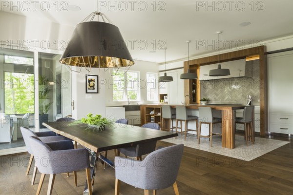 Chrome and walnut wood table with charcoal linen armchairs, large black and gold industrial style pendant lighting fixture and walnut wood island with grey leather and wooden high chairs on travertine mosaic floor surrounded by engineered hickory wood floorboards in dining room kitchen area inside a luxurious contemporary bungalow style home, Quebec, Canada, North America