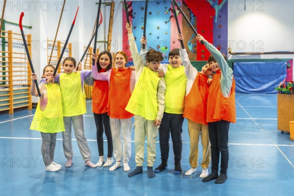 Group of cheerful students wearing colorful bibs raising hockey sticks and celebrating in school gym class