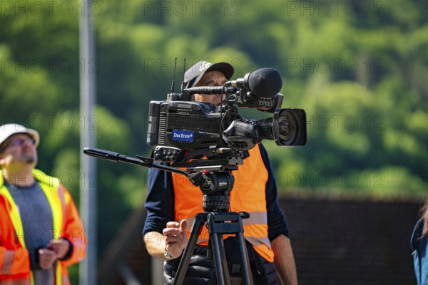 A cameraman in a brightly coloured waistcoat with a camera films outside in sunny weather against a green background, Hermann Hesse Bahn bridge lift, Calw, Germany, Europe