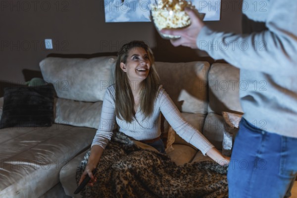 Smiling woman sitting on sofa with remote control, covered with blanket, taking popcorn from boyfriend while watching movie together at home