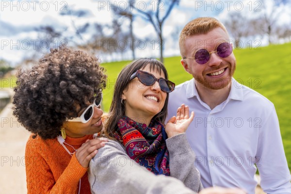 Three cheerful friends, wearing sunglasses, are taking a selfie in a park, enjoying a sunny day and having fun together