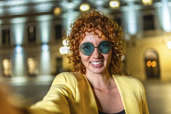 Modern woman with curly hair and sunglasses taking selfie in the city at night