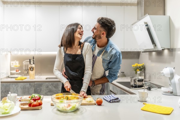 Young loving couple wearing aprons, preparing a healthy salad together in their modern kitchen, embracing and laughing