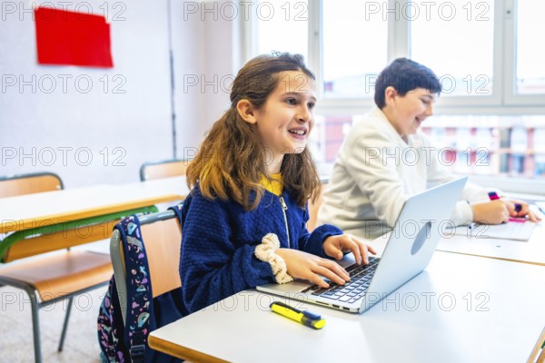 Smiling elementary school girl using laptop during lesson in classroom, next to classmate writing on notebook