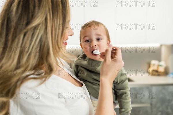 Smiling mother feeding yogurt to her baby son with a spoon in a bright kitchen, healthy eating habits from young age