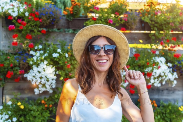 A woman wearing a straw hat and sunglasses is smiling in front of a wall of flowers. The flowers are in various colors and are arranged in a way that creates a beautiful and vibrant display