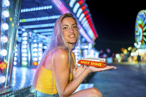 Blonde skinny woman eating a hot dog in an amusement park at night