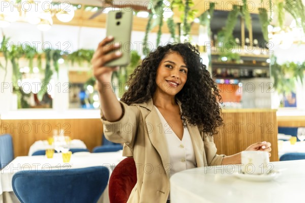 Happy and beauty latin woman with long curly hair and chic clothes taking selfie sitting alone in cafeteria