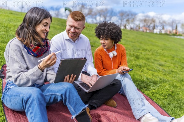 Three university students collaborating on a project, using a digital tablet and laptop while sitting on a blanket in a park
