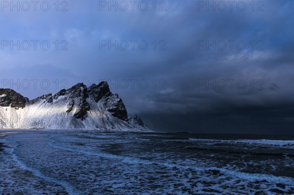 White foamy waves on the beach of Stokkness, behind snowy rocky slopes of Vestrahorn, near Höfn, Sudausturland, Iceland, Europe