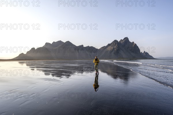 Tourist at black lava beach, sandy beach and sea, mountains Klifatindur reflected in the water, Eystrahorn and Kambhorn, headland Stokksnes, mountain range Klifatindur, Austurland, East Iceland, Iceland, Europe