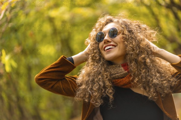 Happy model with curly hair and sunglasses posing and smiling in a park in autumn
