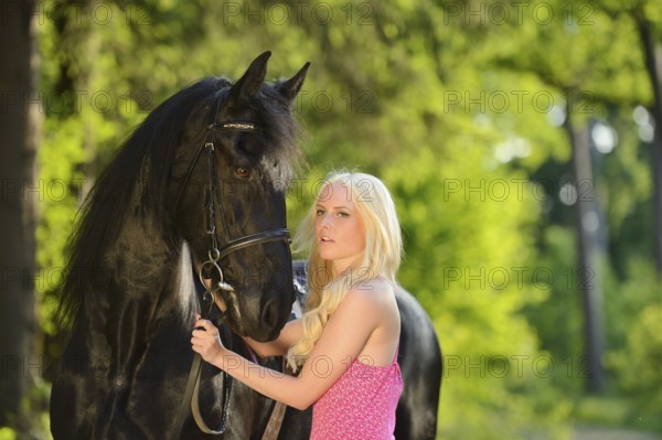 Blonde Woman in a pink dress holding a black Friesian horse on its bridle in a forest setting, Bavaria
