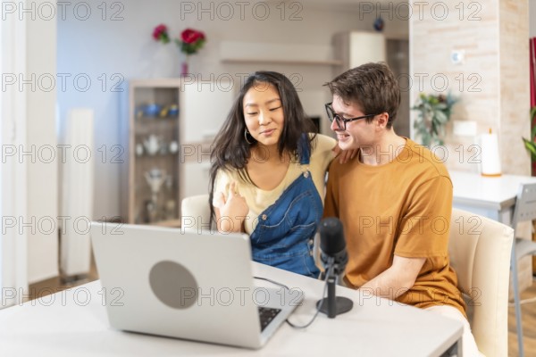Multiethnic young couple using a laptop and a microphone, recording a podcast from their home