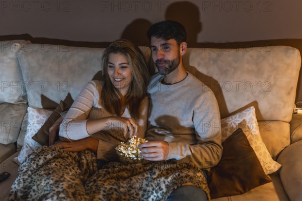 Young couple enjoying a movie night at home, sharing popcorn and relaxing on the sofa under a cozy blanket
