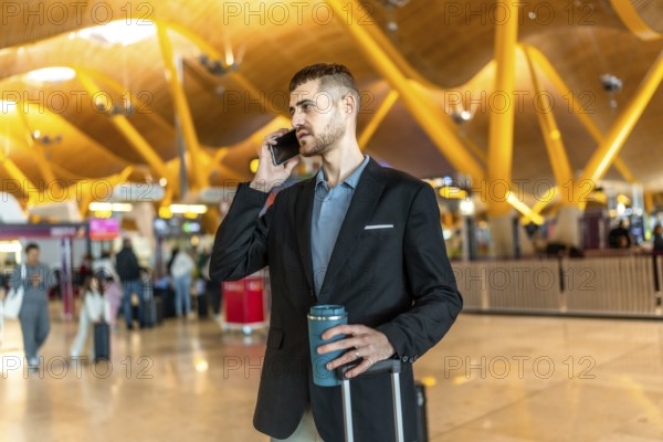 Businessman making a phone call while holding a reusable coffee cup and pulling his luggage at a modern airport terminal