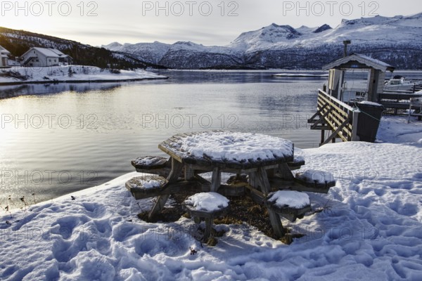 Snow-covered table and benches by the lake with mountains in the background, quiet winter atmosphere, Strávnjunnje, Innhavet, Norway, Europe