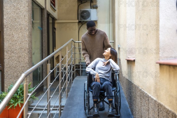 African man helping a disabled work colleague pushing the wheelchair along a ramp next to coworking space building